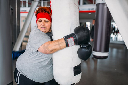 生活艰苦摄影照片_戴手套的超重女子，在健身房拿着拳击袋进行拳击锻炼。卡路里燃烧，肥胖女性在体育俱乐部进行艰苦训练