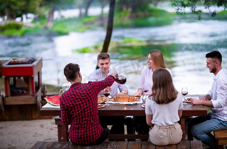 朋友野餐摄影照片_暑假期间，一群快乐的朋友在美丽的自然河边户外野餐法国晚餐