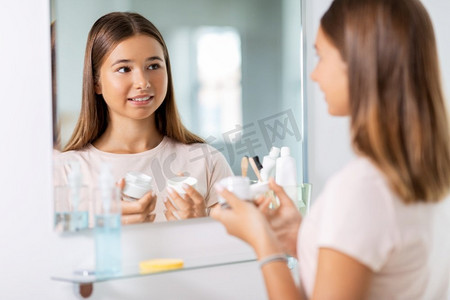 保湿护肤摄影照片_美容、卫生和人的概念—十几岁的女孩与保湿霜看着镜子在浴室。少女与保湿霜在浴室