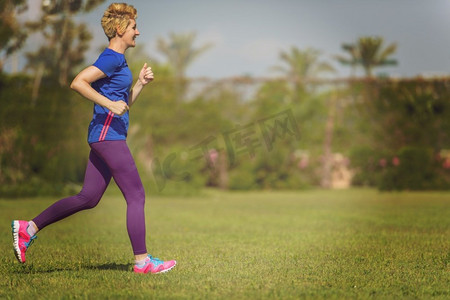 健身运动女摄影照片_阳光明媚的早晨，在美丽的热带公园，年轻的运动女跑步者为马拉松训练