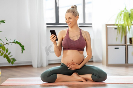 电话应用摄影照片_瑜伽、运动和人的概念快乐孕妇在家中使用智能手机。孕妇拿着智能手机在家做瑜伽