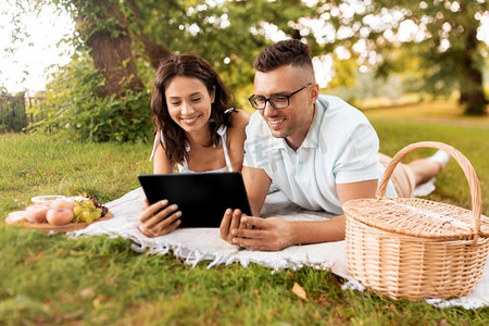 休闲与人的概念快乐的夫妇和平板电脑在夏令营野餐。带着平板电脑在公园野餐的幸福夫妻