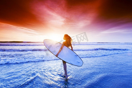 美丽的混血女子在日落时手持冲浪板站在热带海滩上。海滩上的一名女子手持冲浪板