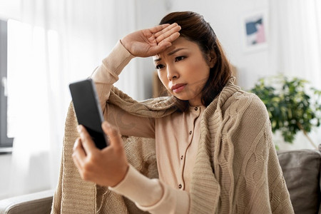 技术，健康和冷概念—悲伤生病的亚洲妇女毯子使用智能手机在家里。悲伤的生病的妇女在毯子使用智能手机在家里