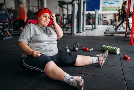 疲惫的胖女人坐在健身房的地板上。卡路里燃烧，肥胖女性在体育俱乐部