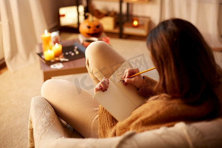 万圣节,假期和休闲概念 — 年轻女子用铅笔写日记,在舒适的家里把脚搁