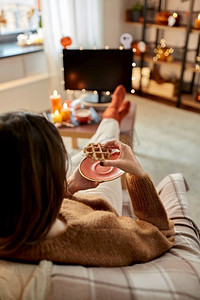万圣节，假日和休闲概念年轻妇女看电视和吃华夫饼在舒适的家里。女人在万圣节看电视吃华夫饼