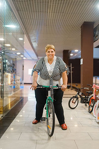 胖女人骑自行车在商场展示。超重的女性在超市里骑自行车，肥胖问题