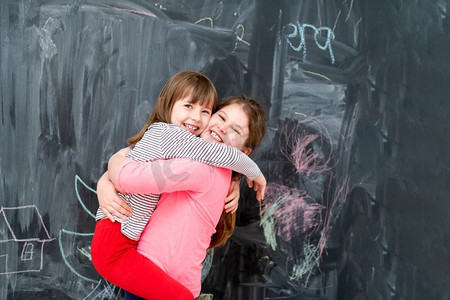 两个快乐可爱的小女孩在黑色黑板前玩耍时互相拥抱的肖像美女女孩