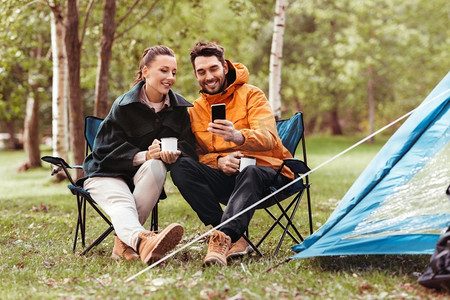 露营、旅游和旅行概念-在露营地点与智能手机喝茶，这是一对幸福的情侣。与智能手机一起在露营地喝茶