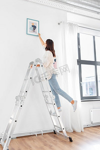 装饰家装海报摄影照片_家装、装修与人的理念--幸福微笑的女人站在梯子上用艺术装饰家。梯子上的女人用艺术装饰家
