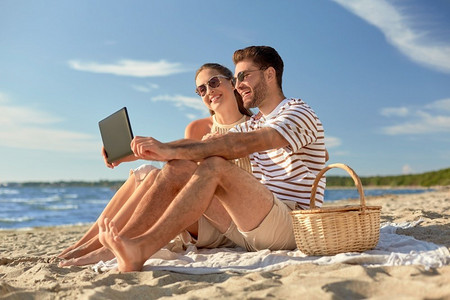 休闲与人的概念-在夏季海滩上，带着平板电脑和野餐篮子的幸福夫妇。带着平板电脑在海滩野餐的幸福夫妻