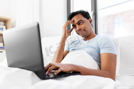 人，技术和休息概念—无聊的印度人有膝上型计算机躺在床上在家里。无聊的印度人与笔记本电脑在床上在家