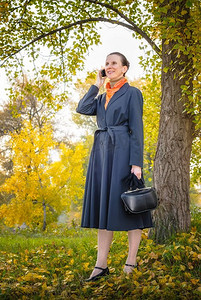 高级商业妇女与移动电话，包和橙色围巾，走在公园，树下在秋天