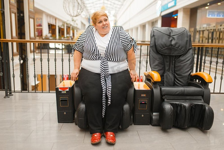 肥胖女性摄影照片_商场里不会把胖女人放在按摩椅上。超市超重女性试图坐在皮革扶手椅上，肥胖问题