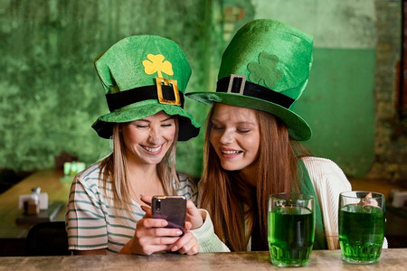 快乐的女性朋友一起庆祝圣帕特里克节与智能手机酒吧
