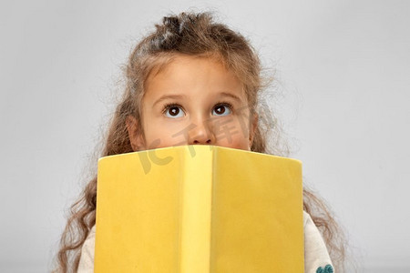 黄皮香瓜摄影照片_阅读、教育和童年观念--小女孩躲在灰色背景下的黄色书本后面。躲在黄皮书后面的小女孩