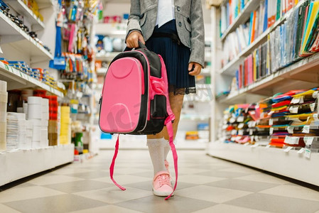 文具店货架上拿着书包的小女生。女孩子在商店买办公用品，学童在超市