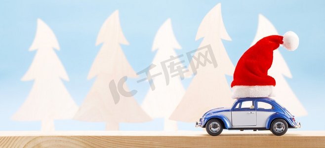 圣诞老人帽子摄影照片_有圣诞节圣诞老人帽子的蓝色玩具汽车在木森林背景，文本的复制空间带圣诞老人帽的玩具车