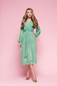 金发碧眼的年轻女子，穿着优雅的青绿色连衣裙。模特在粉色背景上全长摆姿势。珠宝和发型。时尚写真