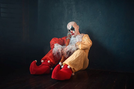 恐怖小丑摄影照片_疲惫不堪的拿着棒球棍的小丑坐在地上。化妆狂欢节装扮的男人，疯狂的疯子