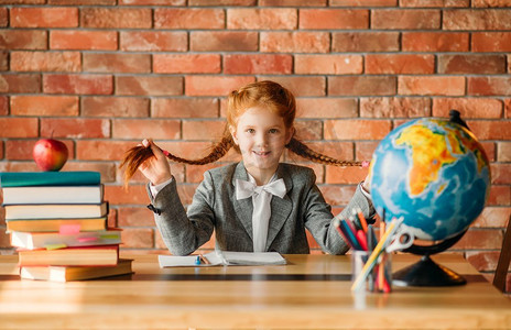 可爱的女学生坐在桌子前，正面视图。女学生在桌子与教科书，苹果和地球仪，做家庭作业的年轻女孩