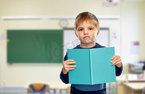 课堂背景摄影照片_学校，教育和学习的概念—不高兴的小男孩在课堂背景下看书。不高兴的小男孩与书在学校