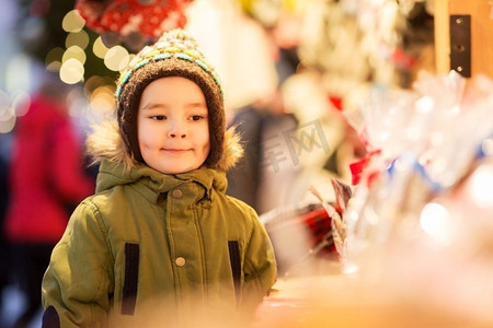 假日，童年和人的概念—快乐的小男孩在圣诞市场摊位在冬天的晚上。快乐的小男孩在圣诞市场在冬天