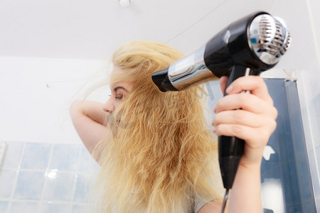 积极的女人使用吹风机在她的金发发型。头发护理，发型设计概念..金发女郎的女人使用吹风机