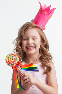 白色小皇冠摄影照片_美丽的小糖果公主，戴着王冠，手里拿着大棒棒糖，笑着。戴皇冠的女孩拿着棒棒糖