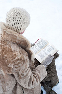 冬天，在一个被白雪覆盖的公园里，一个穿着毛皮大衣、戴着羊毛帽的成年妇女坐在石凳上看书