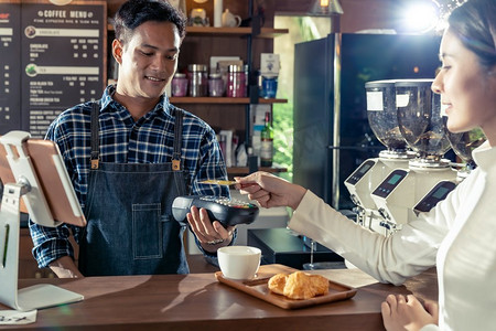 一名亚裔顾客使用非接触式nfs技术的信用卡向咖啡师支付她在咖啡馆购买咖啡的费用。