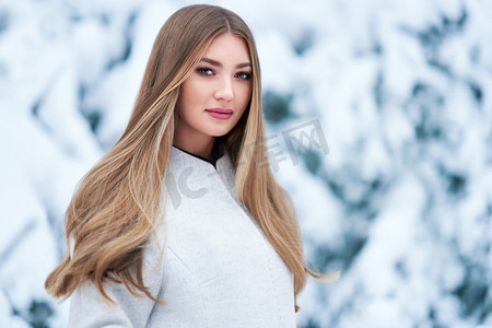 时尚的年轻女子在冬季森林。年轻优雅的模特在时尚的白色外套在性质。金发女郎的女孩与长的光滑的头发
