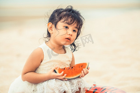 吃女孩西瓜摄影照片_可爱的孩子吃西瓜在热带沙滩在夏天。可爱的孩子在沙滩上吃西瓜。