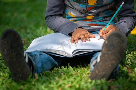 儿童教育画册封面摄影照片_小学生坐在公园的绿草地上，用铅笔在笔记本上写字。儿童教育理念。