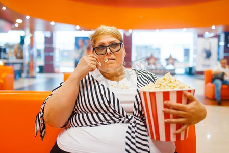 肥胖女性摄影照片_胖女人吃爆米花在电影厅，不健康的垃圾食品。商场里超重的女性，肥胖问题