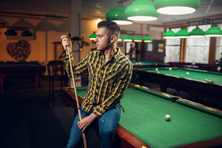 男台球运动员与球杆姿势在绿色桌子。男子在体育酒吧玩美式台球游戏。在桌子上摆出球杆姿势的男性台球运动员