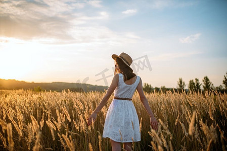 日落时分，一位身着白色连衣裙、头戴草帽的女子走在麦田里。夏日草地上的女性，后景