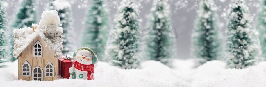 雪人礼物摄影照片_小装饰雪人与礼物在冷杉林附近的木屋在落雪。冬天的雪人和房子