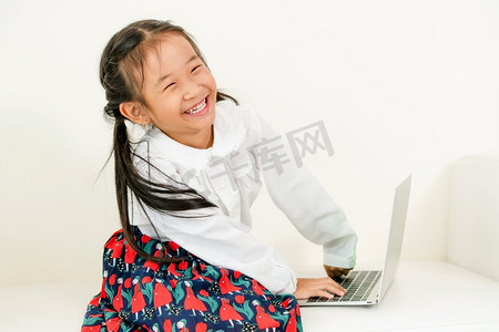 坐在白色沙发上的使用膝上型计算机的小快乐的孩子。童年生活方式使用膝上型计算机的小孩在白色沙发上。