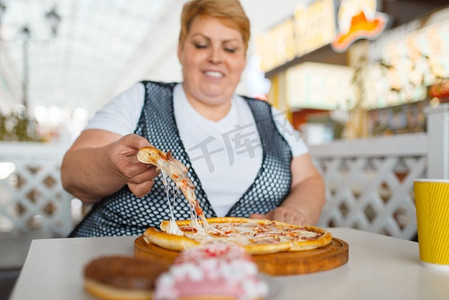 胖女人吃披萨在快餐店，不健康的食物。超重的女性人在桌子上与垃圾晚餐，肥胖问题