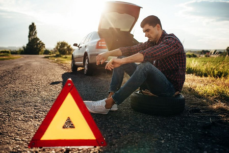 破碎的汽车概念，男人坐在轮胎对破碎三角在沥青路。车辆故障，警告标志