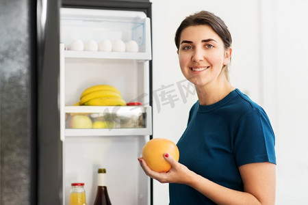 健康的饮食，食物和饮食概念—快乐的女人从冰箱在家庭厨房拿葡萄柚。快乐的女人从家里的冰箱里拿食物