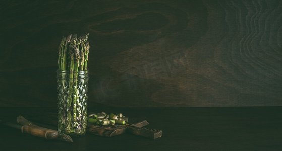 深色墙壁背景摄影照片_玻璃瓶中的绿色芦笋，深色墙壁背景下放在桌子上的水。新鲜芦笋店。春季时令蔬菜。复制空间