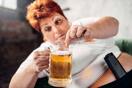 肥胖女性摄影照片_超重的女人喝啤酒吃三明治，暴食症，肥胖。不健康的生活方式，肥胖女性。超重女人喝啤酒，肥胖