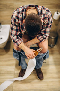 一个男人拿着一瓶啤酒，脱着裤子坐在马桶上，俯瞰着，在家庭聚会之后。醉酒男性头痛患者