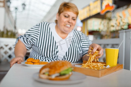 胖女人在商场餐厅吃高热量食物。超重的女性人在桌子上与垃圾晚餐，肥胖问题