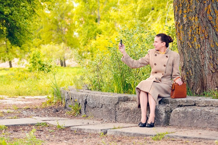 阳光明媚的春日，一位优雅的女商人坐在石墙上，用手机拍照