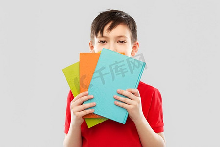 学校，教育和人的概念—小学生男孩在红色t恤隐藏在灰色背景的书籍后面。害羞的学生躲在书本后面