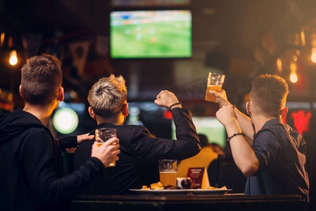 11人制足球场摄影照片_三名男子在体育酒吧看足球电视，球迷陪伴的快乐休闲